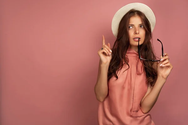 En brunett står på en rosa bakgrund i en stråhatt, ser till toppen, hålla upp ett finger och bita boja av glasögon. — Stockfoto