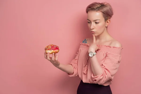 En flicka med kort rosa hår i en blus ser eftertänksamt på en munk i handen, på en rosa bakgrund. — Stockfoto