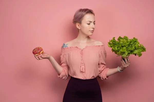 En tjej med kort rosa hår i en blus håller en munk och sallad i händerna, stirrar på det nospartiet, på en rosa bakgrund. — Stockfoto