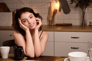 Kıvırcık koyu saçlı bir kız masada elleri ile mutfakta oturur ve onlara kafa dinlenme, gülümseyerek kameraya bakarak