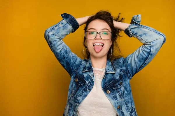 Uma menina encaracolada em jeans e óculos fica em um fundo amarelo, olhando para a câmera com a língua para fora e as mãos nos cabelos. — Fotografia de Stock