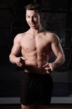 Çıplak gövdeli kaslı bir adam spor salonunda duruyor ve gülümsüyor, işaret parmaklarını öne doğru tutarken çerçeveye bakıyor..