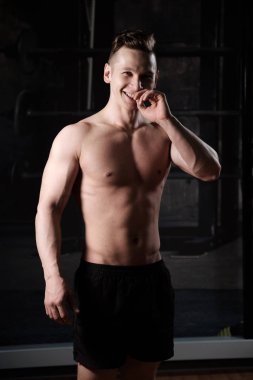 Çıplak gövdeli kaslı bir adam spor salonunda duruyor ve gülerek çerçeve içinde görünüyor.