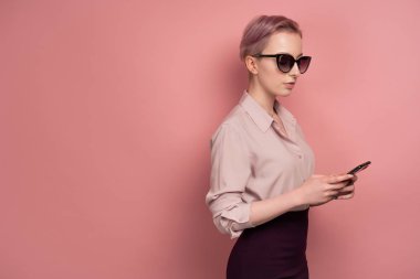 Bir bluz, etek ve koyu gözlük kısa saçlı bir kız pembe bir arka plan üzerinde yarım dönüş duruyor ve akıllı telefon bakar
