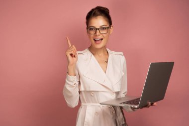 Hafif ceket ve gözlüklü bir kız dizüstü bilgisayar tutar ve işaret parmağını en üste çıkararak kameraya sevinçle gülümser..