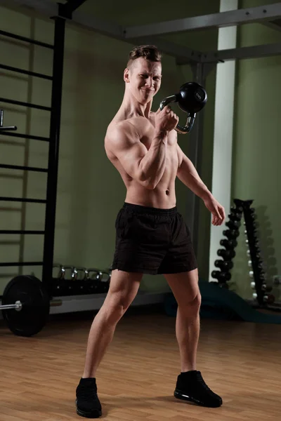 Un tipo musculoso con un torso desnudo en pantalones cortos deportivos está ejecutando un kettlebell con una mano con una cara apretada . —  Fotos de Stock