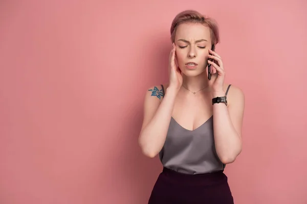 Egy lány, rövid rózsaszín haja és a tetoválás egy szürke felső becsukta a szemét feszülten, beszél a telefon, a rózsaszín háttér. — Stock Fotó