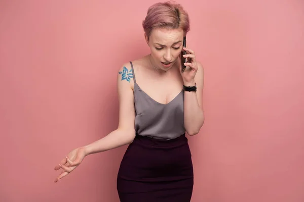 짧은 분홍색 머리와 회색 상단에 문신을 가진 소녀는 분홍색 배경에 서있는 동안 무의식적으로 전화로 말합니다.. — 스톡 사진