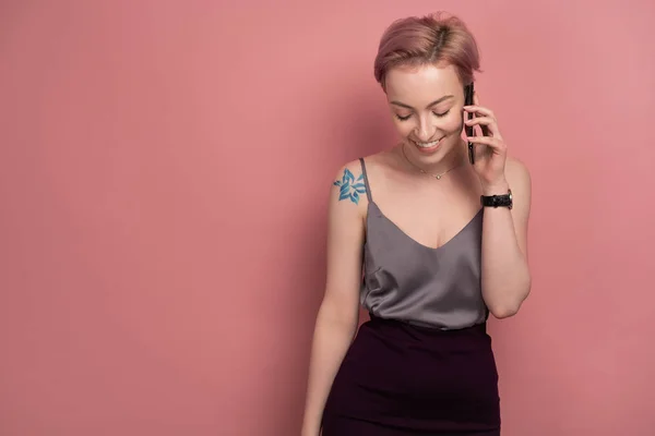 Una chica con el pelo corto de color rosa y un tatuaje en una parte superior gris habla vergonzosamente por teléfono mientras está de pie sobre un fondo rosa . — Foto de Stock