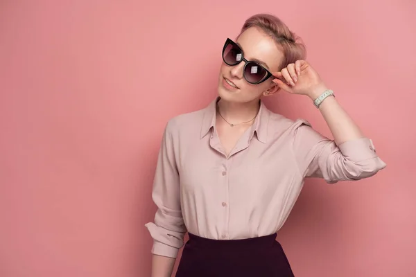 En tjej med kort rosa hår i en blus och kjol, leende, tittar på kameran i solglasögon, stående på en rosa bakgrund. — Stockfoto
