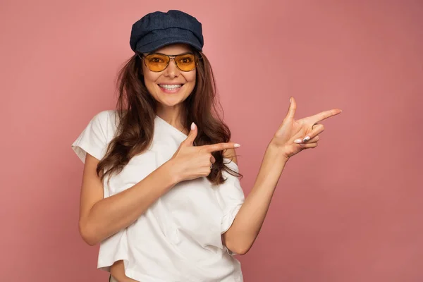 Eine Brünette in weißem T-Shirt, blauer Mütze und Sonnenbrille lächelt breit und zeigt mit den Fingern zur Seite auf rosa Hintergrund — Stockfoto