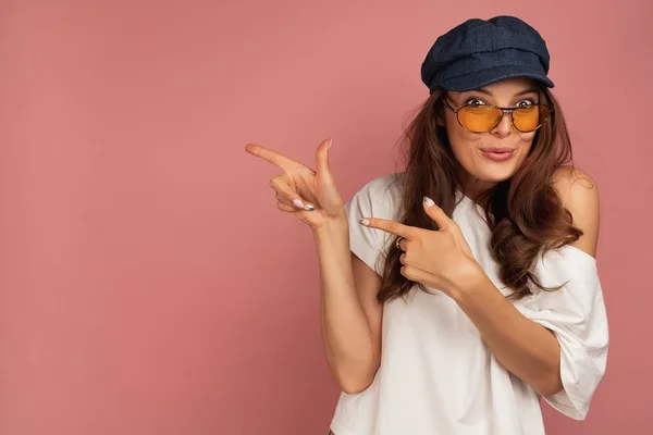 Morena em uma camiseta branca, boné azul e óculos de sol com um rosto engraçado fica em um fundo rosa e aponta os dedos para o lado — Fotografia de Stock