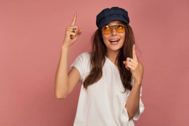 Beyaz bir t-shirt, mavi şapka ve güneş gözlüğü bir esmer parmaklarını işaret eder ve üst gözleri ile görünüyor.