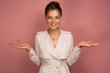 Bir esmer parlak bir trençkot içinde pembe bir arka plan üzerinde duruyor saçlarını ayrı, yanlara kollarını yayarak, avuç içi kadar.