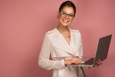 Açık ceket ve gözlük lü koyu saçlı bir kız pembe bir arka plan üzerinde elinde bir dizüstü bilgisayar ile duruyor ve kameraya gülümsüyor.