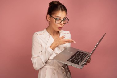 Bir esmer toplanan saç ile pembe bir arka plan üzerinde duruyor ve bir dizüstü bilgisayar işaret ederken şaşkınlıkla kameraya bakar.