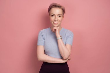 Bir t-shirt kısa pembe saçlı bir kız, kameraya bakarak ve eliyle çenesini propping, pembe bir arka plan üzerinde duruyor.