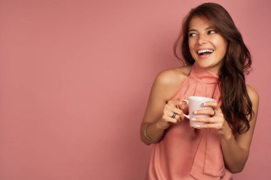 Elinde bir kupa ile pembe elbiseli koyu saçlı bir kız pembe bir arka plan üzerinde duruyor ve sevinçle yan gülüyor görünüyor.