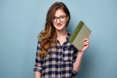 Ekose elbise ve gözlüklü genç bir kız elinde bir kitap ile mavi bir arka plan üzerinde duruyor ve kameraya bakar.