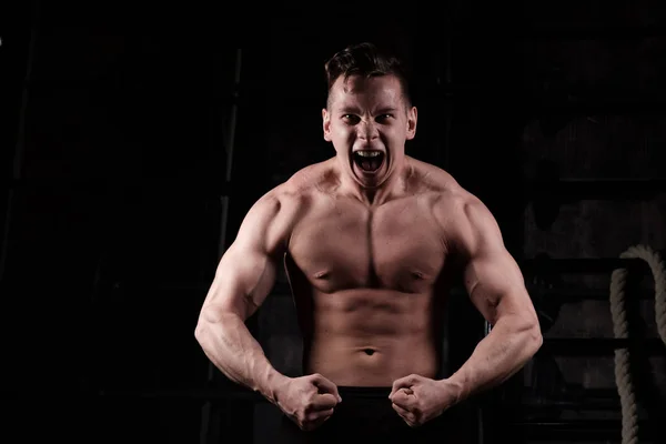 Un tipo inflado con un torso desnudo se para en el gimnasio tensando sus músculos y gritando, mirando dentro del marco . — Foto de Stock