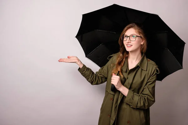 Una chica con una capa verde y gafas está de pie sobre un fondo blanco con un paraguas negro y con la mano abierta mira hacia arriba . — Foto de Stock