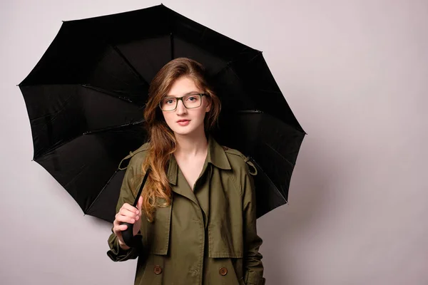 Egy lány egy zöld kabátot és üvegek áll a fehér háttér egy fekete esernyő, és nézi a kamerát. — Stock Fotó