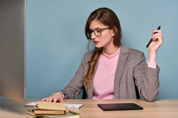 분홍색 스웨터, 회색 재킷과 안경을 쓴 소녀가 컴퓨터에 앉아 손에 펜을 들고 모니터를 바라보고 있습니다.. — 스톡 사진