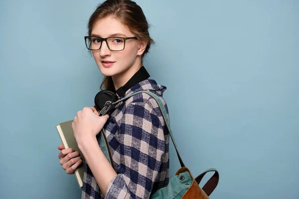 격자무늬 셔츠와 안경을 쓴 소녀가 목에 헤드폰을 쓰고 책과 배낭이 있는 파란색 배경에 서 있습니다.. — 스톡 사진