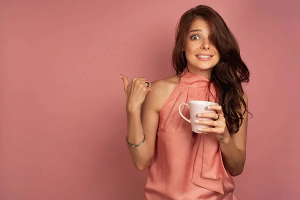 Una chica con un top rosa está de pie sobre un fondo rosa con una taza en la mano sonriendo falsamente y mostrando un pulgar hacia atrás . — Foto de Stock