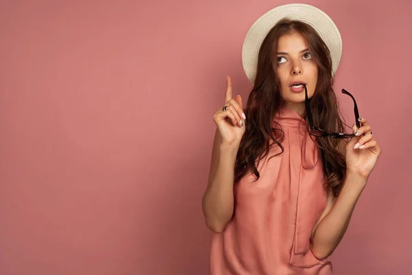 Menina de cabelos escuros em um vestido rosa e um chapéu de palha fica em um fundo rosa e olha atentamente e aponta um dedo para o topo — Fotografia de Stock