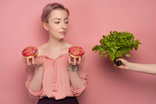 A girl with short hair, holds donuts in her hands and raising an eyebrow, looks at her hand with a salad, on a pink background. — Stock Photo, Image