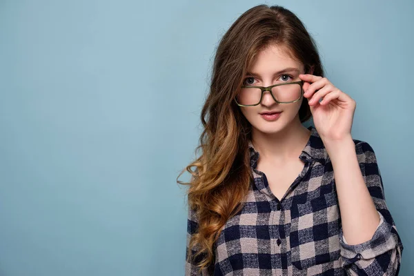 En ung flicka i en pläd klänning står på en blå bakgrund och tittar på kameran över glasögon. — Stockfoto