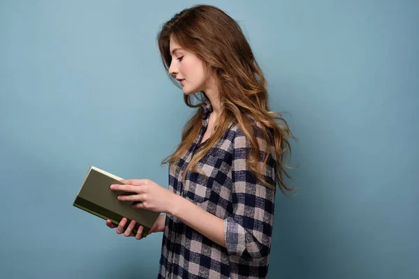 En ung flicka i en pläd skjorta står i profil på en blå bakgrund och ser ner på boken med huvudet böjt. — Stockfoto