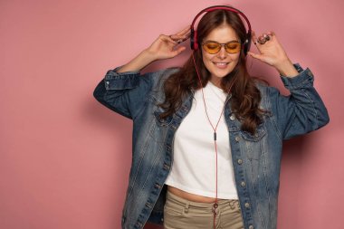 Kot pantolonlu bir kız, gözleri kapalı ve dudağını ısırarak kulaklık ve gözlüklerle pembe bir arka plan üzerinde duruyor müzik dinliyor.