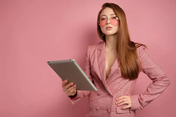 Rödhårig flicka i en jacka står på en rosa bakgrund med en elektronisk tablett och tittar in i kameran över glasögon. — Stockfoto