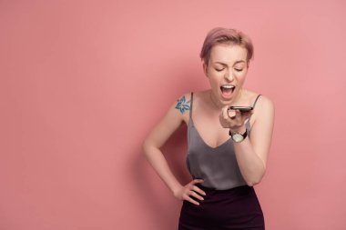 Kısa pembe saçlı ve omzunda gri bir bluz olan bir kız, pembe bir arka plan üzerinde dururken telefona bağırır..