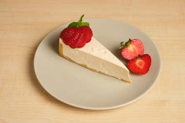 Hafif ahşap bir masa üzerinde çilek dekore cheesecake ile bir tabak duruyor.