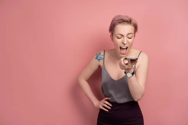 En tjej med kort rosa hår och en tatuering på axeln i en grå topp skriker på telefonen medan du står på en rosa bakgrund. — Stockfoto