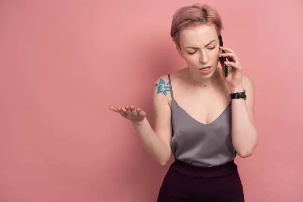 Una chica con el pelo corto de color rosa y un tatuaje en una parte superior gris habla desagradablemente por teléfono, de pie sobre un fondo rosa . — Foto de Stock