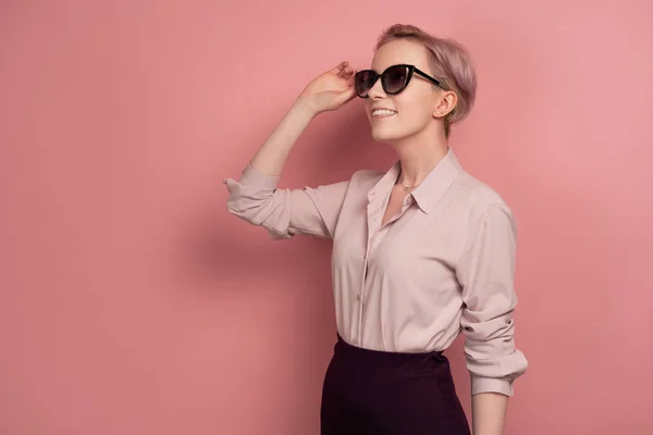 Uma menina com cabelo rosa curto em uma blusa, saia e óculos fica em meia volta em um fundo rosa e olha para o topo . — Fotografia de Stock