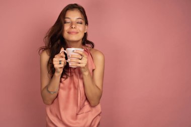 Pembe elbiseli koyu saçlı bir kız elinde bir fincan ile duruyor ve zevk gözlerini kapatarak, aroması teneffüs.