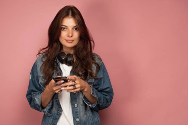 Kot pantolonlu koyu saçlı bir kız, boynunda kulaklıklar ve telefon la pembe bir arka plan üzerinde duruyor ve kameraya bakıyor..