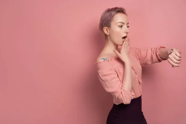 Ein Mädchen mit kurzen Haaren in einer Bluse mit nackten Schultern schaut schockiert auf ihre Uhr, im Profil vor rosa Hintergrund stehend — Stockfoto