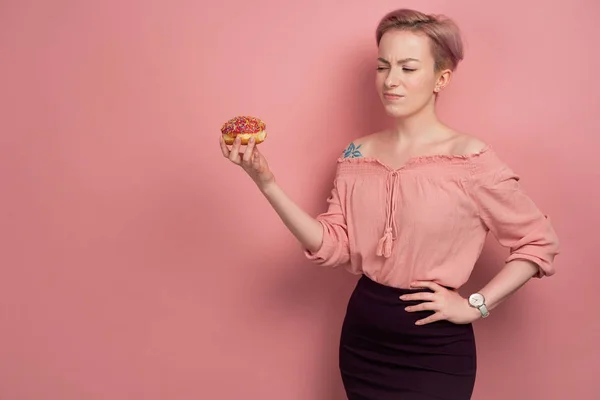 짧은 분홍색 머리를 가진 소녀, 그녀의 손에 도넛을 잡고, 분홍색 배경에, 그에게 얼굴을 찡그린다. — 스톡 사진