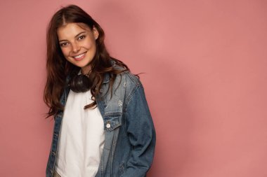 Esmer boynunda kulaklık ile kot pantolon pembe bir arka plan üzerinde sevinçle gülümseyerek yarım dönüş ayakta kameraya bakar.