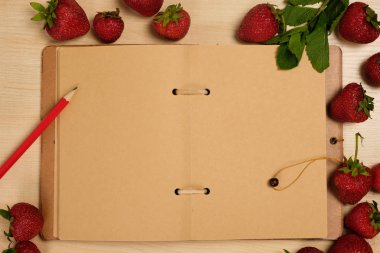 Kırmızı kalemli açık bir zanaat kağıt defteri, hafif ahşap bir masanın üzerinde çileklerle çevrilidir..