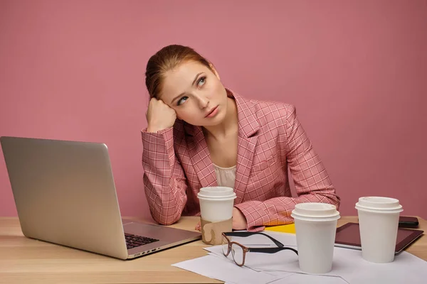 Rothaariges Mädchen in Jacke, das an einem Tisch vor einem Laptop auf rosa Hintergrund sitzt, mit dem Kopf in der Hand und nach oben schauend. — Stockfoto