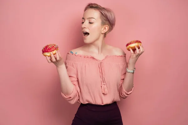 En flicka med kort rosa hår, håller munkar och öppnar munnen, når för en av dem, på en rosa bakgrund. — Stockfoto