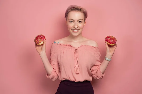블라우스에 짧은 분홍색 머리를 가진 소녀는 분홍색 배경에, 카메라에 즐겁게 찾고, 그녀의 손에 도넛을 보유. — 스톡 사진