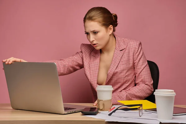 Ruiva em um terno de negócios em um fundo rosa sentado em uma mesa com papéis e café e olhando tensamente para um laptop . — Fotografia de Stock